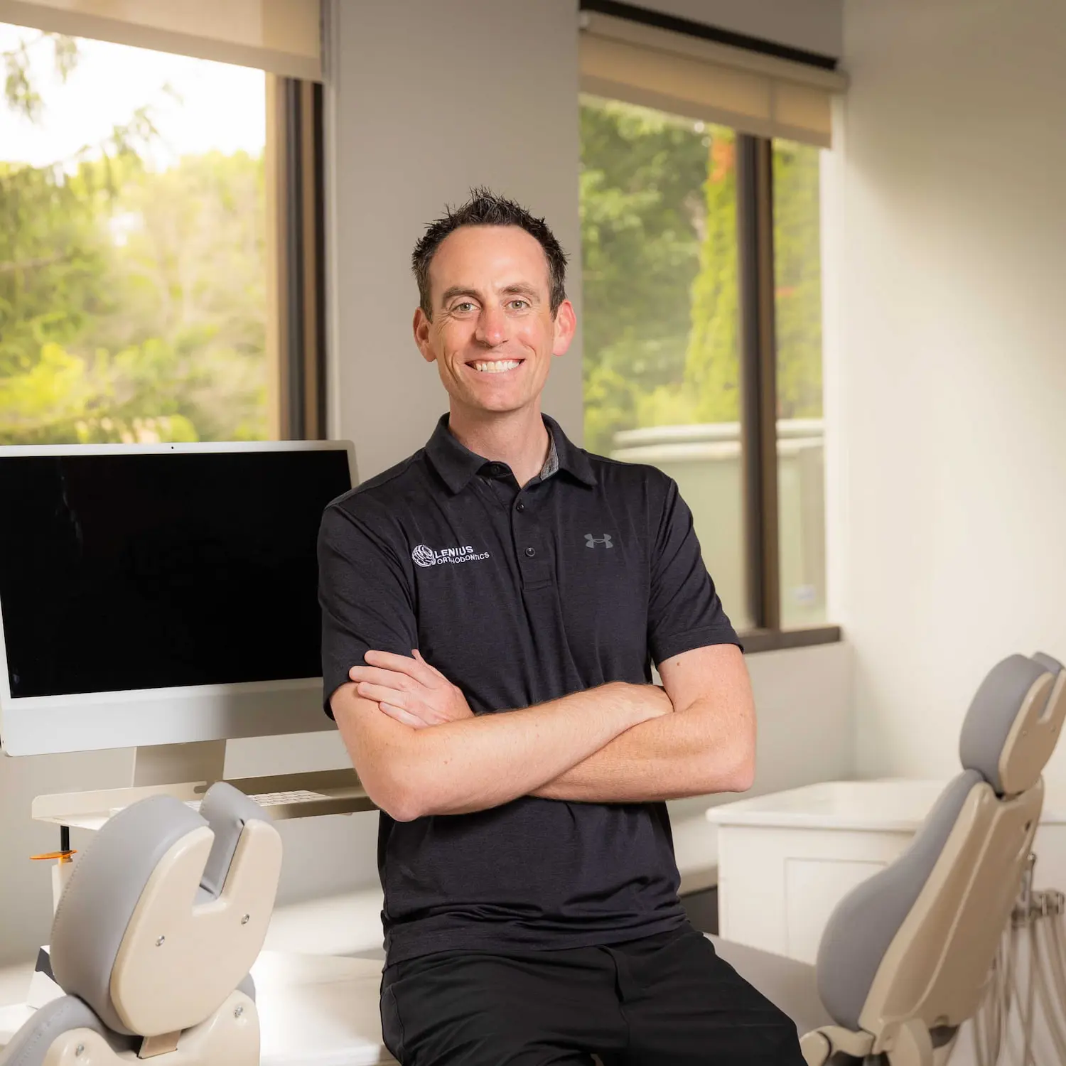 Smiling man in black polo stands with arms crossed represent All Orthodontic Insurance Benefits at Lenius Orthodontics in Overland Park, KS, surrounded by dental chairs and greenery.