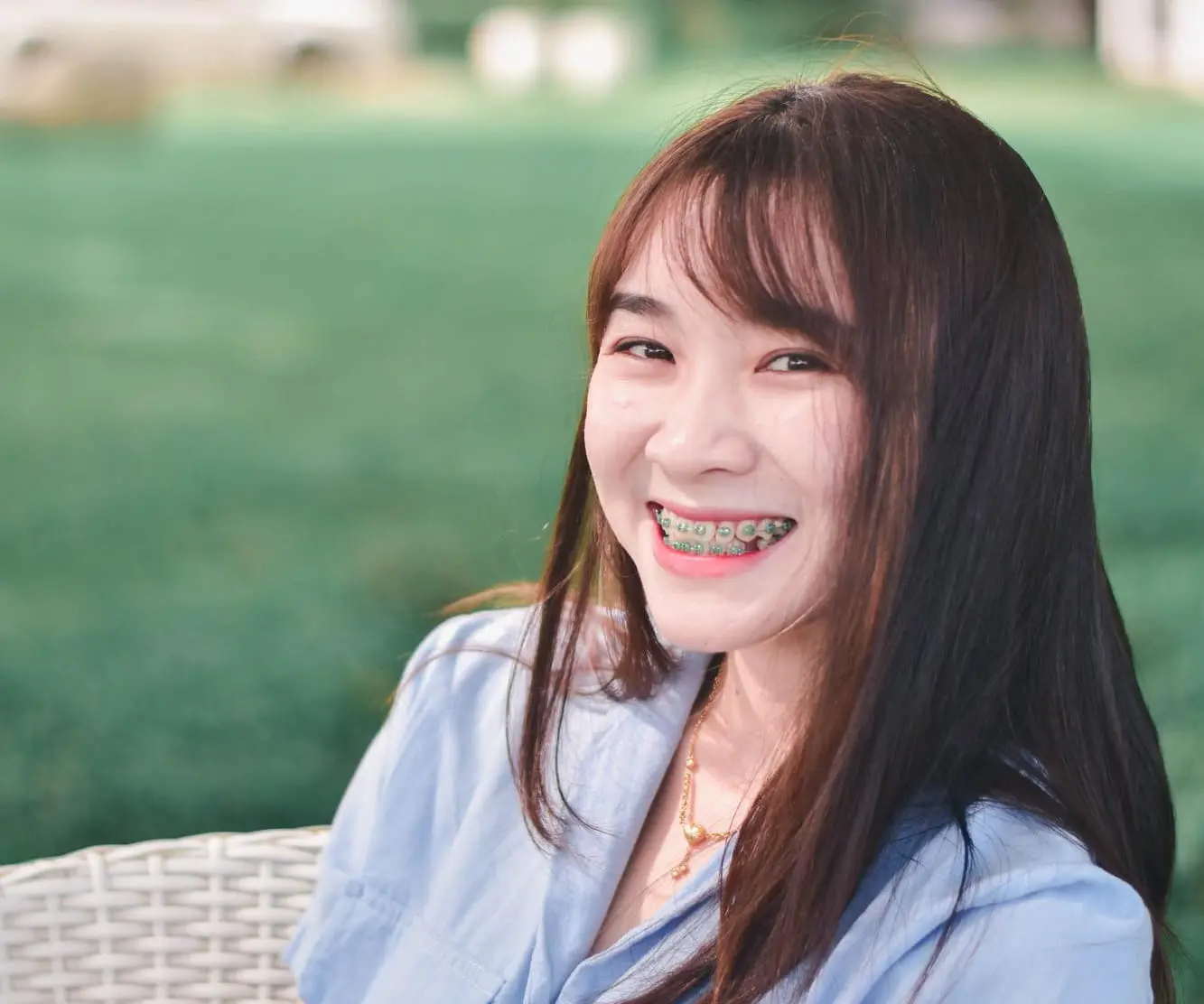 A young woman with braces smiles outdoors on a wicker chair with green lawn at Lenius Orthodontics in Overland Park, KS.