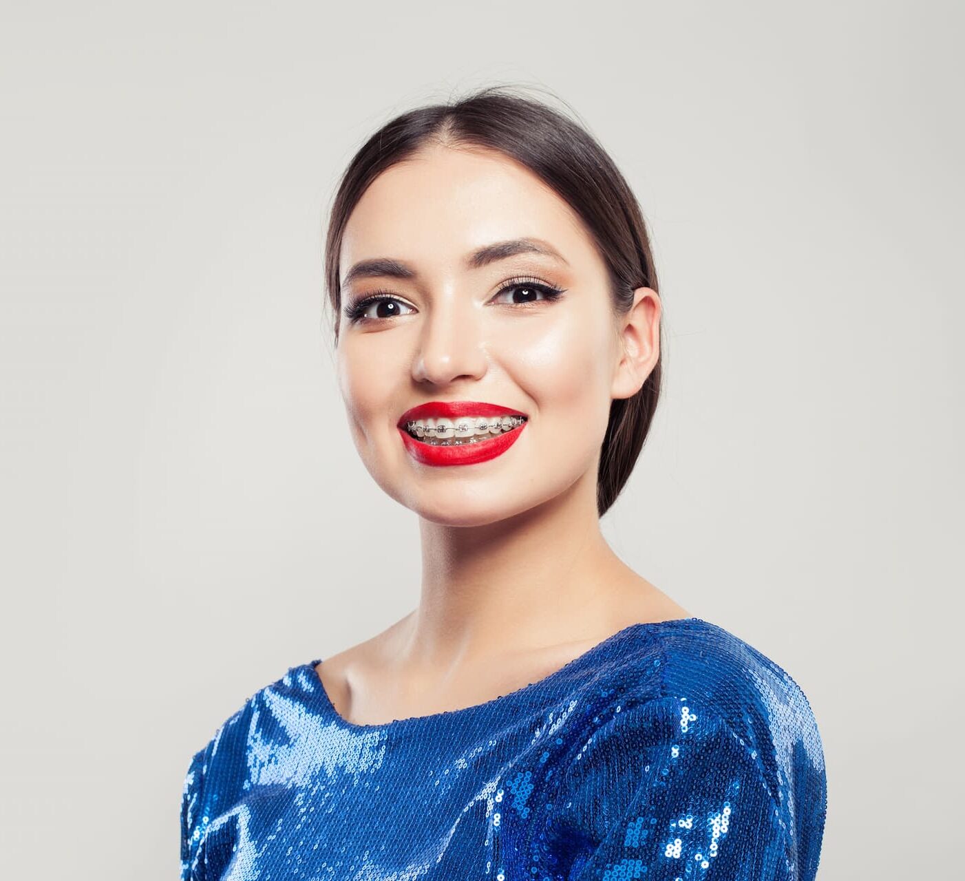A confident woman in a blue sequin top smiles, revealing adult braces from Lenius Orthodontics in Overland Park, KS.